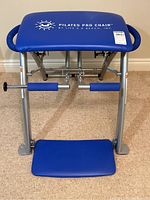 Front view of the Pilates Pro Chair showing blue padded seat with logo and aluminum frame with springs and foot pedal.