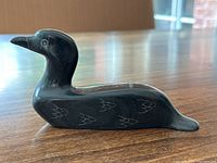 Side angle of black soapstone bird sculpture showing smooth carving, engraved feather detail, and artist signature visible on bottom.