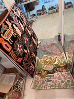 Two needlework rugs displayed leaning against a sliding mirrored closet door, showing vibrant and pastel embroidery designs with birds and floral themes.