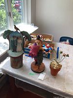 Wood carved sculpture with man, woman, stylized trees and bird on stump base placed on table among other items.