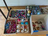 Overview photo showing four boxes containing various vintage and novelty Christmas ornaments including colorful garland beads, assorted glass bulbs, and Old World Christmas novelty ornaments.
