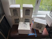 Six boxes of Department 56 Shops of Dickens' Village houses shown stacked on a small wood table near window. Each box has a black and white illustration of a different house.