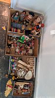 Three cardboard boxes filled with assorted vintage dolls and figurines laid on floor against wall. Boxes contain dolls dressed in various cultural costumes and styles.