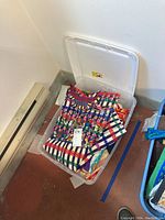 Plastic storage bin on floor filled with folded textiles, top shows colorful embroidered piece