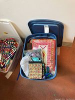 Top-down view of blue plastic storage bin filled with assorted folded textiles including multi-colored fabrics, printed panel, pouch and braided cord