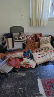 Wide view showing multiple woven blankets, embroidered pillows, a fabric handbag, and various textiles arranged on a chair and on the floor.