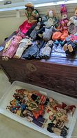 Wide view showing multiple dolls and marionettes arranged on a carved wooden chest and in a plastic bin below