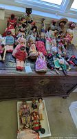 Wide view of multiple multicultural themed cloth and composite dolls including Caribbean style and other ethnic dolls, arranged on a wooden chest and in a white tray below revealing various designs and costumes.