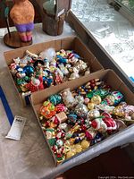 Two cardboard boxes containing a large assortment of vintage Christmas tree ornaments of various shapes, colors, and designs, laid out on a table next to glassware.