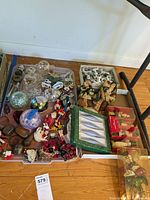 Wide view of various Christmas ornaments including nutcrackers, balls, figurines, bread figure, and boxed fish-shaped ornaments on trays seen from above on wooden floor.
