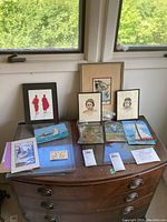 Overview of various framed and unframed art pieces placed on wooden chest below a window with green foliage outside