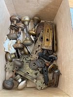 Box of vintage door hardware including several ornate brass and bronze door knobs, backplates with detailed engravings, and lock parts, all with visible wear.