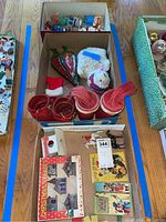 Three cardboard boxes containing the lot's contents: top box with colorful ornament figures and small village set; middle box with red holiday boots, plush bear, leaf ornaments, and small stocking; bottom box with boxed Christmas village set and assorted miniatures.
