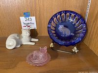 Wide photo showing all three items: blue glass cat bowl with metal stand, white Lenox cat figurine, and pink glass cat pill box.