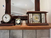 Four mantel clocks on wooden shelf: Bulova, Ansonia, Howard Miller, and quartz cat clock together showing size and style