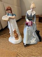 Two Royal Doulton figurines displayed side by side on wood floor showing Childhood Days with girl and orange tabby cat and Dorothy knitting with black cat