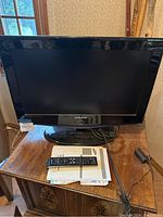 Front view of Samsung 26-inch TV with remote on top of user manual and papers on wooden cabinet.