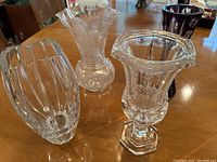 Photo showing all four vases together on a wooden table, including a clear crystal etched vase, ribbed rectangular vase, flared ruffled rim vase, and purple vase with cutouts.