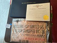 Top-down photo showing black folio embossed The San Francisco Call, certificate of authentication on cream paper with gold seal, and vintage newspaper with headline about missing boy hunted as Folsom kidnapper victim inside plastic sleeve.
