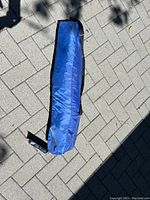 Blue Quick Shade half tent shelter in carry bag laid on paved stone ground under sunlight.