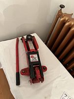 Full view of the red hydraulic floor car jack with handle and wheels, placed on white surface near radiator.