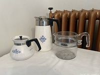 Three coffee pots arranged on a white surface with a copper radiator background.