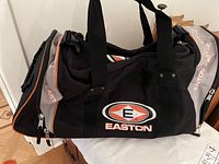 Front and side view of black and gray Easton sports duffle bag with large white and orange logo on side panel, showing zippered compartments and black handles.