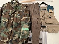 US Army camouflage jacket, brown tweed hunting pants, and Columbia fishing vest displayed on hangers.
