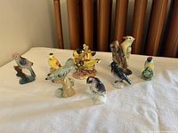 Photo showing 10 various colored ceramic bird figurines arranged in two rows on a table, with backgrounds including a wooden chair back.