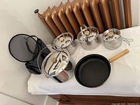 Overview of the camping cooking set on a white surface: three stainless steel pots with lids, one frying pan with wooden handle, and a carry bag.