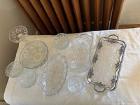 Top down view of nine clear crystal glassware pieces arranged on fabric surface, including a serving tray with silver trim and various bowls and plates of different shapes and sizes.