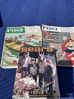 Three vintage magazines laid flat on a blue padded surface. Visible covers show two Saturday Evening Post magazines from the 1950s and one 1983 Sears Fall Winter catalog.