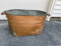 Side view of the antique copper wash tub basin showing copper exterior with dents, verdigris, and metal handles.