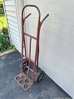 Full side view of the vintage metal hand truck dolly in red, standing upright outdoors on asphalt beside a building and garden.