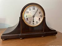 Front view of Kienzle tambour clock showing round white dial with black numerals and wooden curved case base.