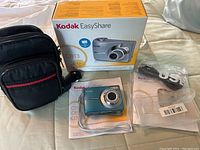 Kodak digital camera with case, USB cord, dock, box, and manuals laid out on bed.