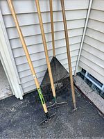 Four long-handled lawn and garden tools leaning against siding, including rakes and one edger.