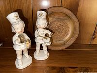 Two matching Japanese ceramic figurines of young band members, one playing cymbals, one playing drum, displayed in front of a carved wooden plate.