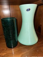 Photo of two Murano glass vases on wooden shelf. One dark green cylindrical vase with crackled texture, one light turquoise flared vase with Murano sticker.