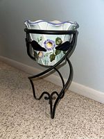 Ceramic plant pot in floral design held by black metal stand with curved legs and leaf decorations.