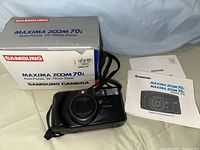Samsung Maxima Zoom 70i camera resting on its original box with black wrist strap visible. The box shows Samsung branding and model details including the price sticker $149.99.