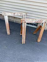 Pair of wooden saw horses shown from a side angle on asphalt, leaning slightly, with visible wood grain and metal brackets.