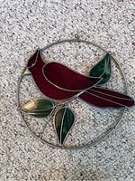 Circular stained glass cardinal ornament laying flat on carpet showing red bird and green leaves within metal circle