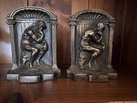 Pair of bronze bookends featuring The Thinker sculpture in a seated pose within shelled niches, placed on wooden shelf background.