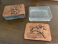 Two vintage trinket boxes, each with copper relief art tops (one floral, one equestrian) and clear glass rectangular bases with geometric patterning.