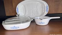 Photo showing all three CorningWare pieces: roasting platter, lidded casserole, and saucepan with handle, all white with blue cornflower floral pattern.