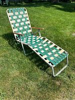 Full view of the vintage aluminum lawn lounge chair on green grass, showing the length and wooden armrest detail with woven strap seating.