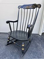 Front angle view of vintage Nichols & Stone black rocking chair showing floral painted accents on top backrest, black spindle back, curved arms and seat, and gold detailing on legs and rockers.