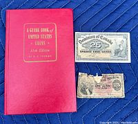 Red hardcover coin guide book and two vintage 25 cent notes, one Canadian and one US, displayed on a blue fabric background.