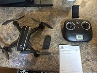 Drone with foldable propellers, camera on front, extra battery next to it, and remote controller with LCD screen.